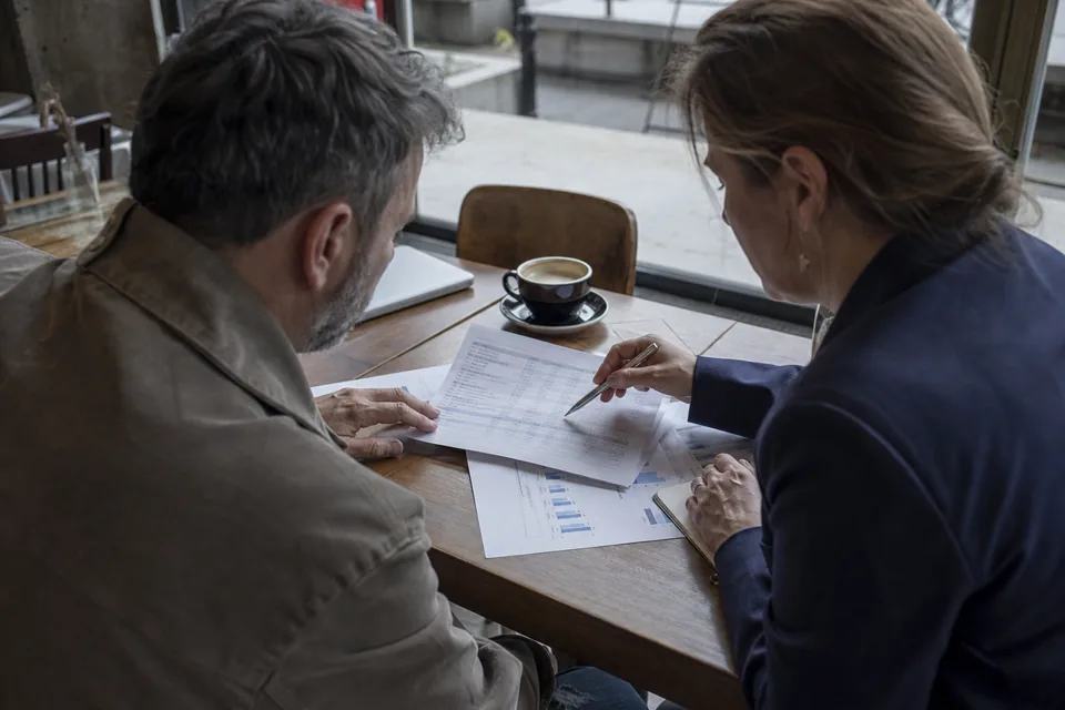 Buyer and seller reviewing printed revenue tables with a pen at a cafe table, seen over the shoulder