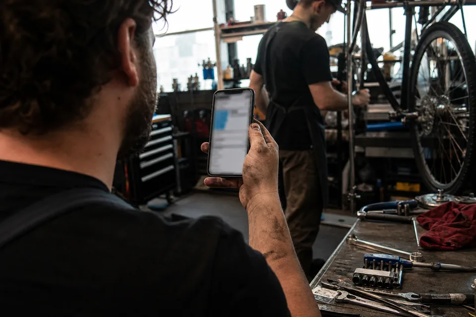 Hand holding a smartphone with an app open in a bicycle repair workshop, tools and wheels in the background