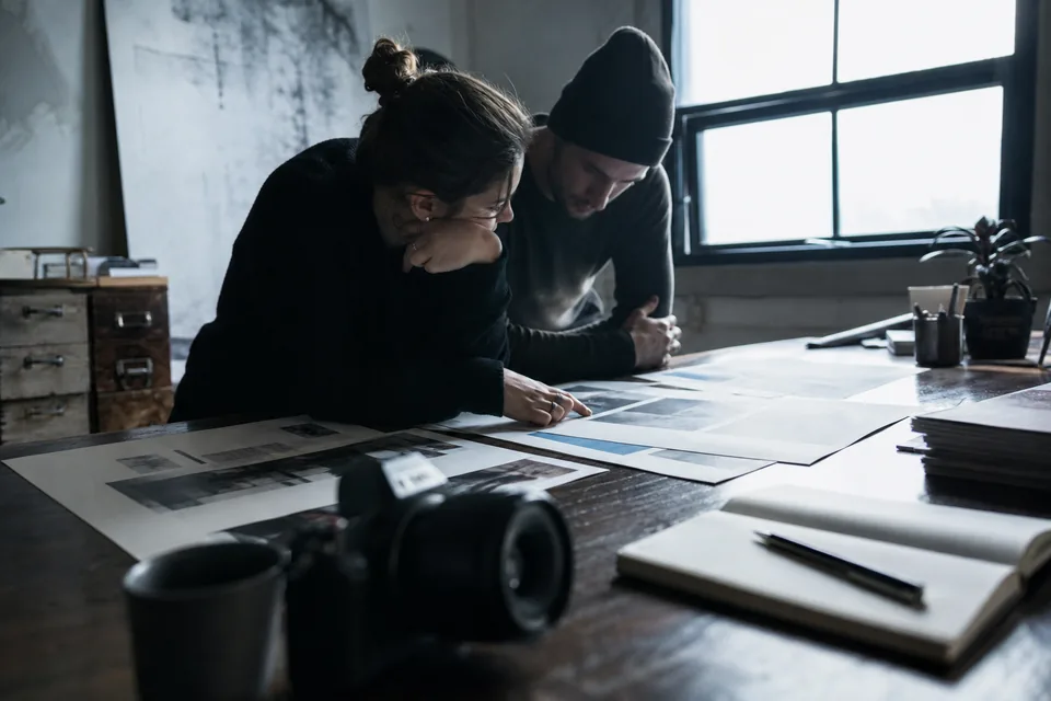 Desk of a content publisher with printed article layouts, a camera and a notebook in morning window light, shot from a low angle