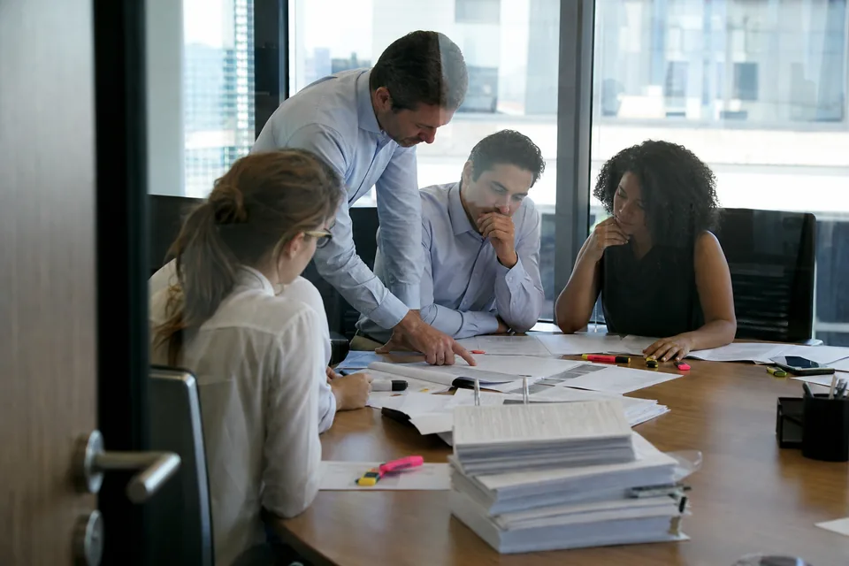Corp dev team around a conference table with printed due diligence pages, photographed through a glass door