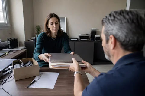 Founder handing a folder of printed credentials and a laptop to a new owner across a desk in a small office